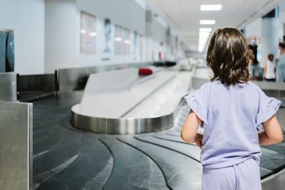 Back view of unrecognizable girl in lilac clothes waiting for luggage to arrive on carousel in airport terminal during trip