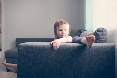 European toddler child sits and gets bored on gray sofa in bright living room, minimalist interior