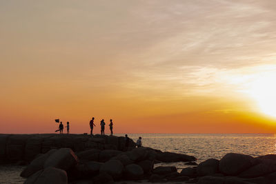 People on rocks by sea against sky during sunset