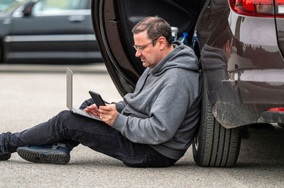 Middle-aged man sitting on the asphalt by the car and working on a laptop, working remotely, travel