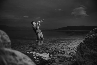 Man playing trumpet while standing in sea against sky at dusk