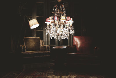 Illuminated chandelier hanging by armchairs in darkroom