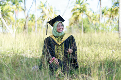 Smiling young woman wearing graduation gown while standing on field