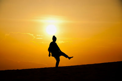 Silhouette woman standing on one leg at beach against orange sky during sunset