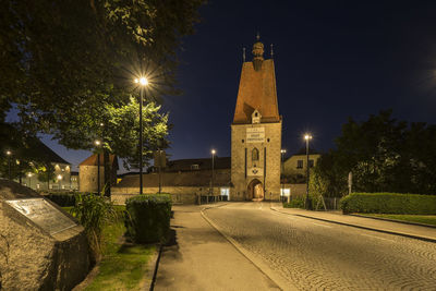 Illuminated street amidst buildings against sky at night