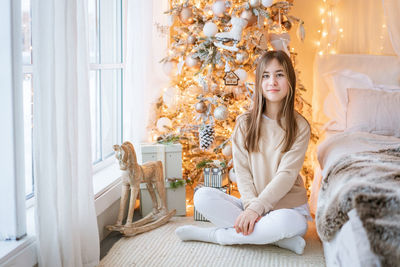 Cute girl sits on floor by window with christmas tree in background in her