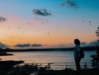 Side view of woman looking at silhouette birds flying over sea against sky during sunset