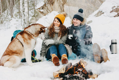 Full length of couple with dog sitting by campfire on snow covered field