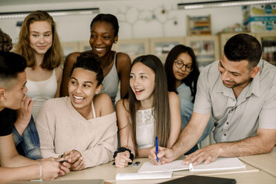 Smiling students looking at teenage boy while teacher teaching over table in classroom