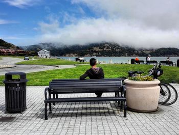 Rear view of man sitting on bench over cobblestone by grass field against sky