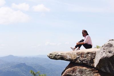 Young woman sitting on cliff against sky