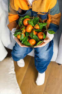 Boy in a bright blue-orange checkered shirt and jeans holds a copper bowl of fresh tangerines.