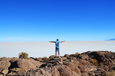 Rear view of man standing on rock against blue sky at desert