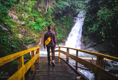 Rear view of man walking on footbridge by waterfall in forest