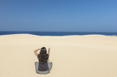 Rear view of woman sitting on sand at beach against clear sky