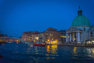 Illuminated buildings at waterfront against blue sky