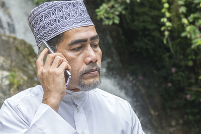 Thoughtful man talking on phone while sitting against waterfall