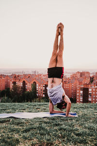 Full length of man with arms raised doing handstand over mat on grass against clear sky