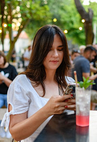 Young woman using mobile phone, drinking cocktail in open air bar in park in city