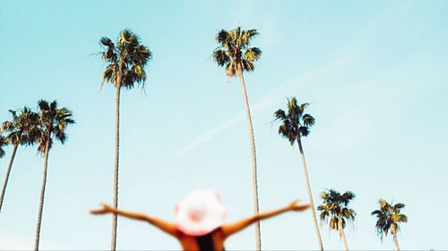 Rear view of woman with arms outstretched standing against palm trees and sky