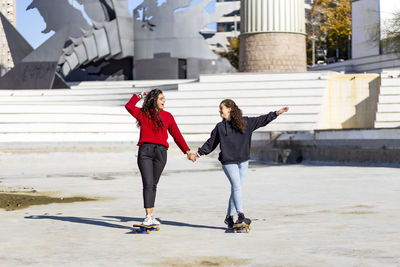 Happy female friends holding hands while skateboarding in city