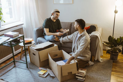 Smiling man sitting on sofa talking to boyfriend unboxing at home