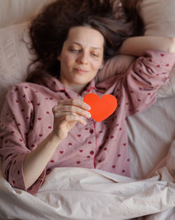 Portrait of young woman holding heart shape on bed at home