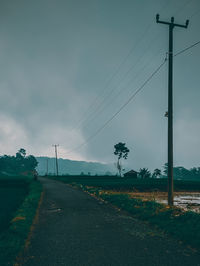 Road by electricity pylons on field against sky