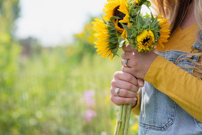 Midsection of young woman in overalls holding yellow sunflowers in field