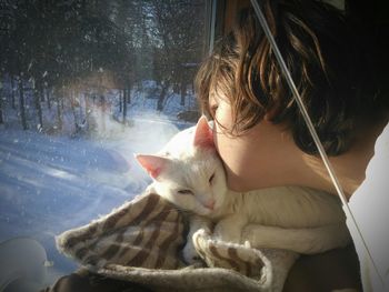 Close-up of girl kissing cat while looking through window during winter