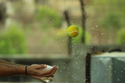 Close-up of hands holding plastic by tennis ball amidst water drops