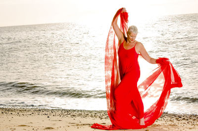 Full length of mature woman wearing red dress with scarf standing on sea shore at beach