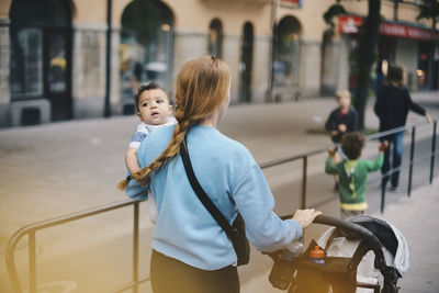 Rear view of mother walking with sons while pushing baby stroller on sidewalk in city