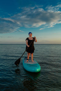 Rear view of woman surfing in sea against sky during sunset