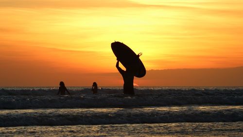 Silhouette surfers standing on beach against sky during sunset