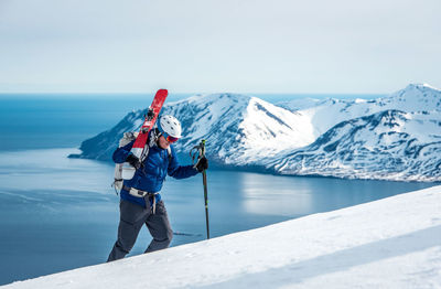 Man hiking uphill with skis and mountains and ocean behind him