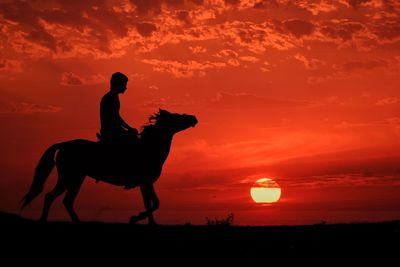 Silhouette man riding horse on field against orange sky