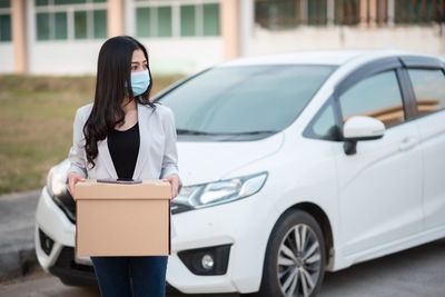 Woman wearing mask holding cardboard box standing by car