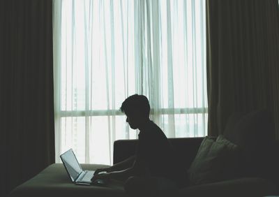 Side view of silhouette young man using laptop while sitting on sofa at home