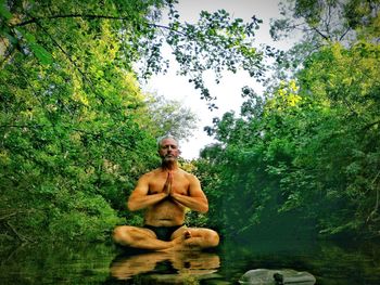 Portrait of shirtless man sitting in forest against sky