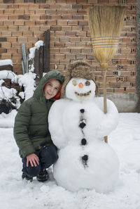 Full length of smiling boy crouching by snowman in yard during winter
