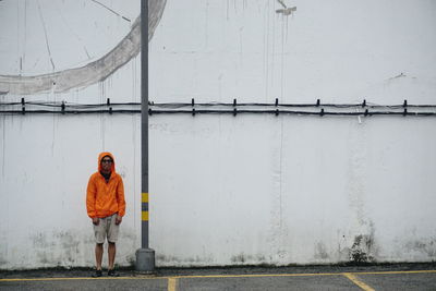 Young man in orange hooded jacket standing against wall