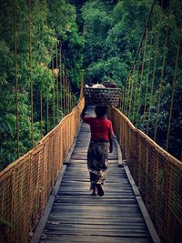 Rear view of woman carrying basket on head while walking on footbridge in forest