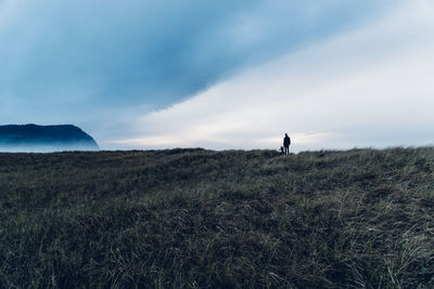 Distant view of person with child on grassy field against cloudy sky