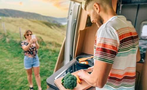 Young man cooking vegetables in a camper van while his wife takes a picture with her cell phone