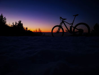 Silhouette bicycle parked on snow covered land against sky during sunset