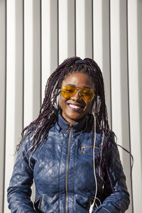Happy young african american female with afro braids dressed in blue jacket and stylish sunglasses enjoying music through earphones while chilling in sunlight against striped wall
