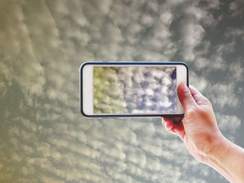 Low angle view of hand holding mobile phone against cloudy sky