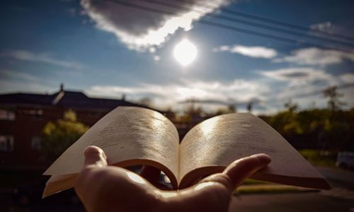 Close-up of hand holding book against sky