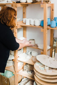 Side view of unrecognizable woman in casual black outfit standing near shelves with various pots and bowls with plates and different pottery while putting small clay vase in light studio